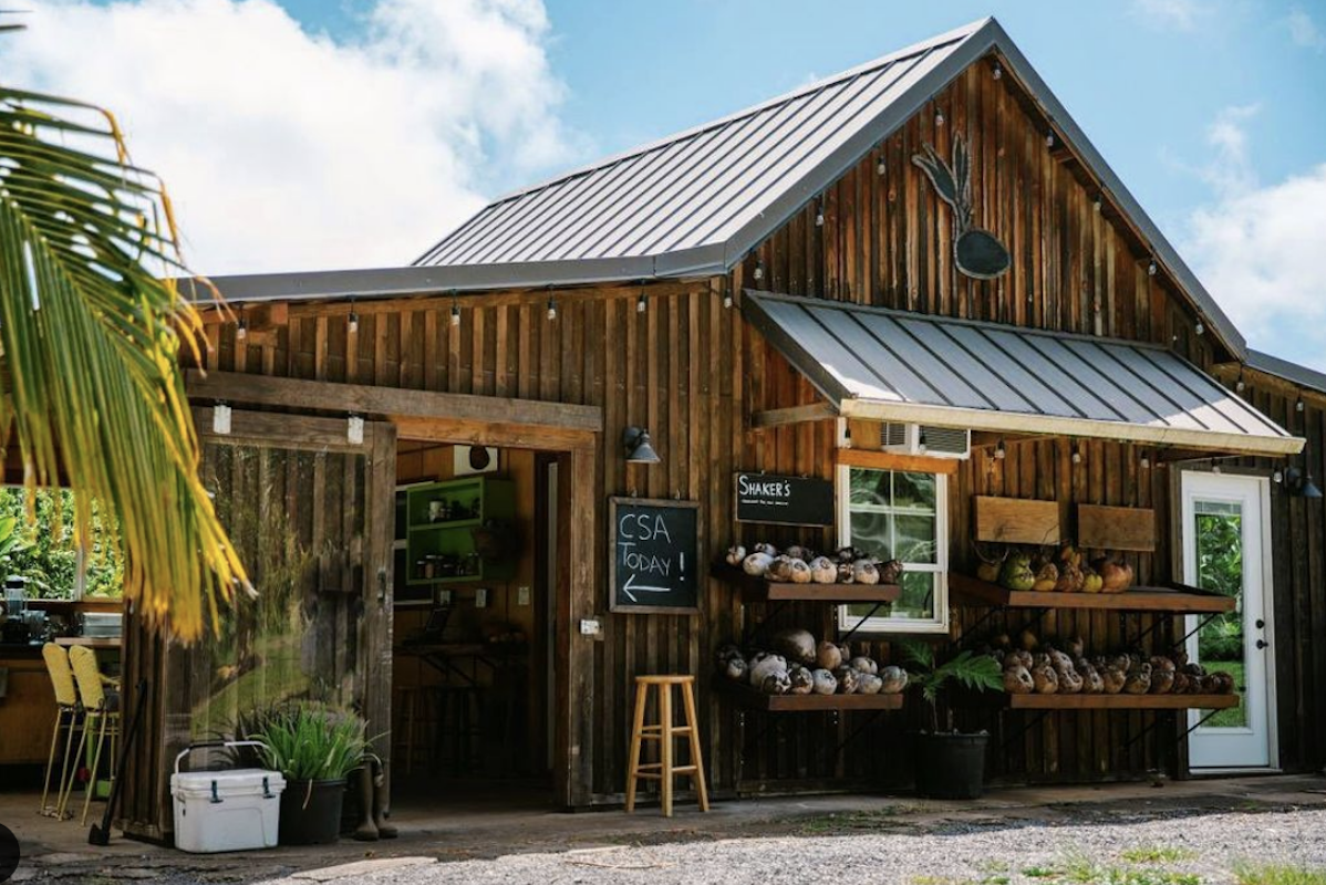 A brown wooden barn from the outside wit shelves of fresh produce outside and string lights hung on the trim.