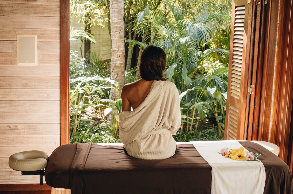 A person with long hair sits on a massage table wrapped in a towel at the best spa hotel in Maui, facing a lush garden with various tropical plants and trees. A window on both sides frames the relaxing scene, and there are flowers placed on the table beside the person.