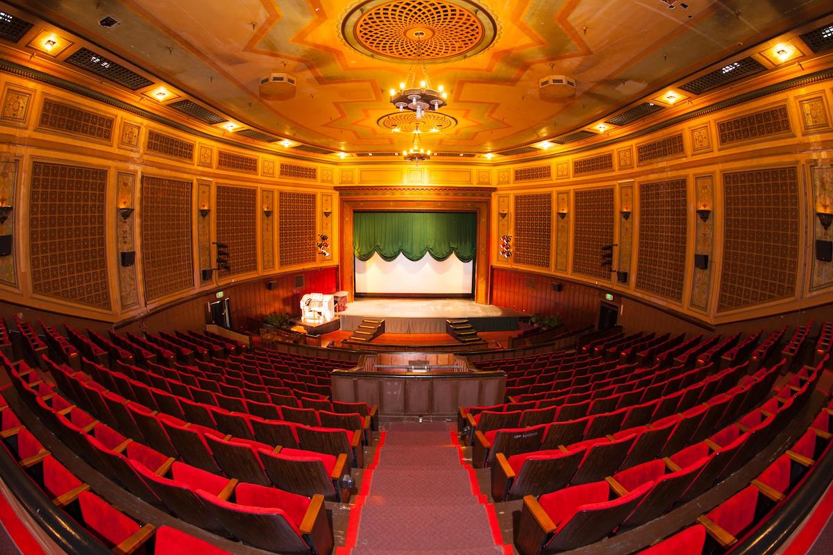 A fish eye lens of a theater with red seats and walkways and orange, lit walls and ceiling and a stage with green curtains.
