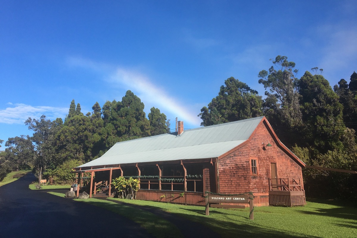 A reddish building with the sign saying "Volcano Art Center" amidst greenery and a blue sky with a rainbow in it.