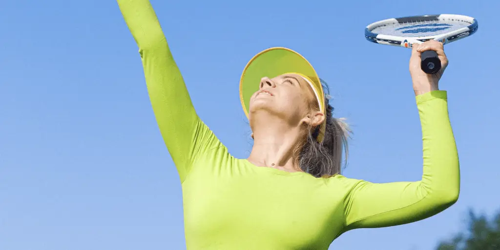 A tennis player in a yellow visor and green long-sleeve UV top for women is preparing to serve, extending an arm upward while holding a tennis racket in the other hand. The sky is clear and blue in the background.