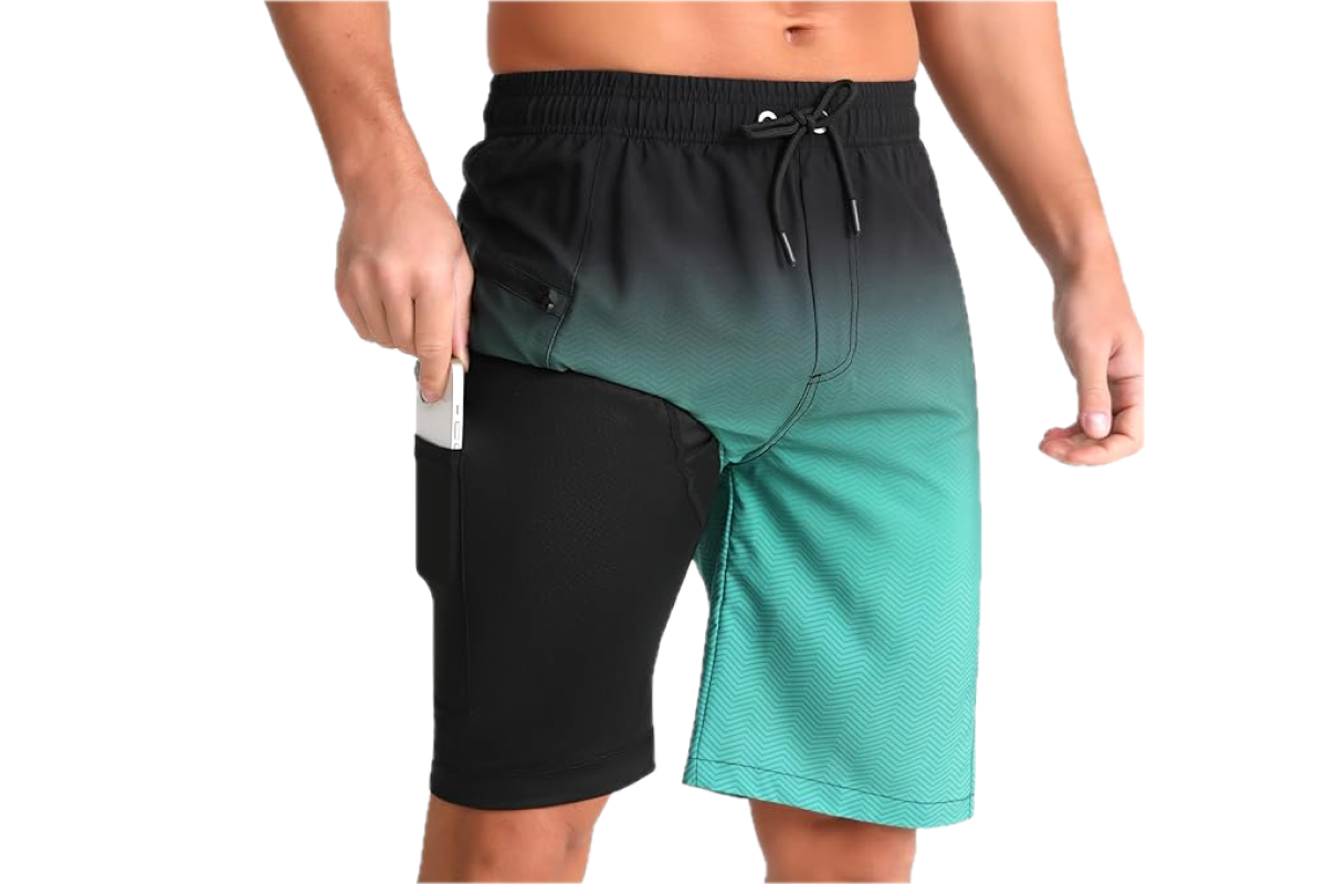 The lower body of a man wearing nothing but a pair of multi colored swim trunks, including black, green and grey, with a white cellphone in the side pocket.