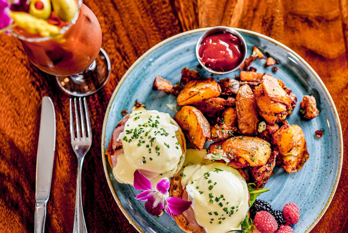 A blue plate with potatoes, eggs benedict, fruit, ketchup and a cocktail on the side.