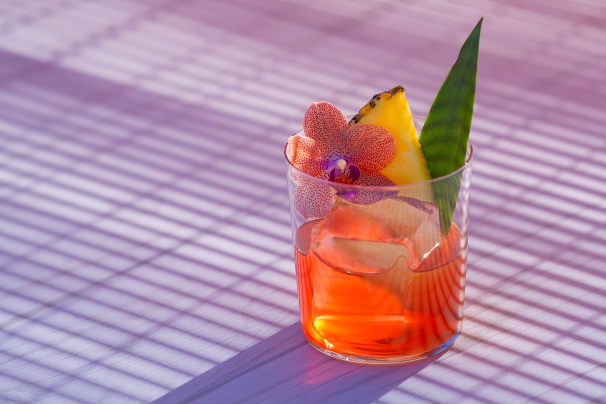 A reddish drink topped with a decorative orchid flower, pineapple slice and leaf.