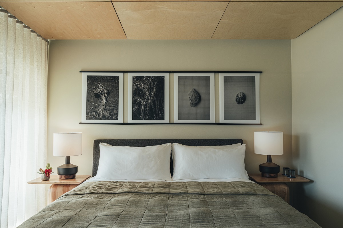 A minimalist bedroom in one of the best vetted value hotels on the Big Island features a neatly made bed with white pillows and an olive-green quilt. Twin modern bedside tables each hold a lamp. Above the bed, a set of four monochrome abstract art pieces hang on the wall. Light flows in through a sheer curtain.