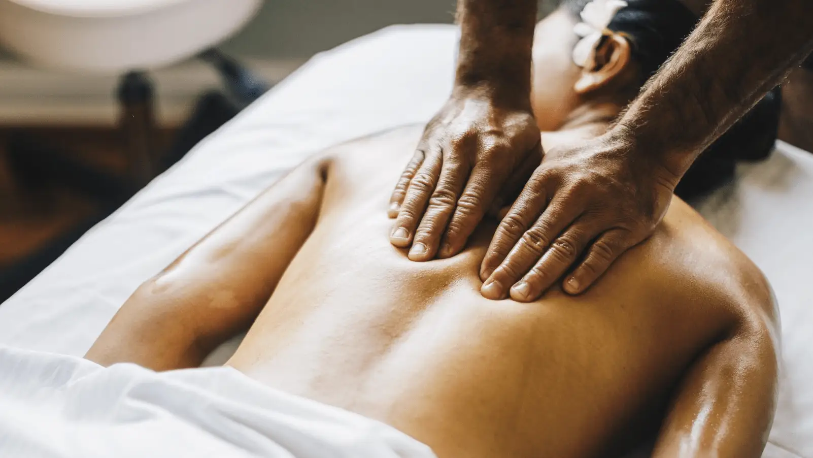 A person is lying face down on a massage table with a white towel covering their lower body. Their upper body is exposed, and a masseuse's hands, possibly using Hawaiian beauty products, are applying pressure to their upper back, providing a therapeutic massage. The atmosphere appears calm and relaxing.
