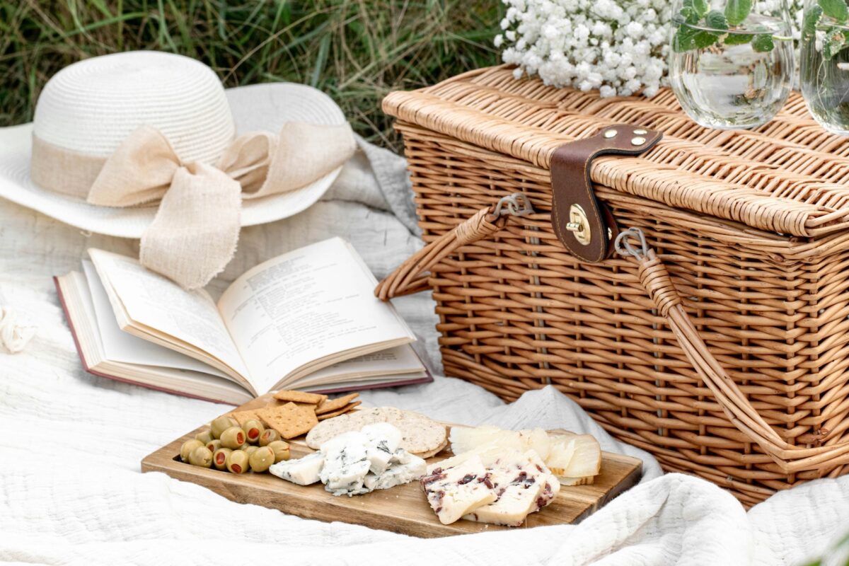 A wicker picnic basket sits on a white blanket, surrounded by greenery and white flowers. Nearby, a wooden board holds an assortment of cheeses and rolled Italian appetizers. Compostable, planet-friendly picnic essentials elevate the experience, while a white summer hat and open book add to the leisurely setting.