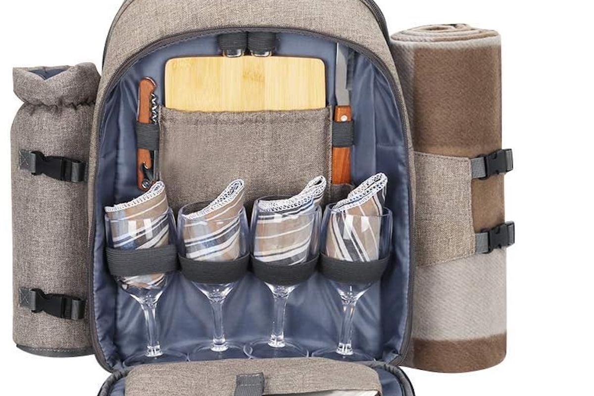 A front-on photo of an open backpack with a rolled blanket attached to the side, wine glasses with napkins, a cutting board and some cutlery.