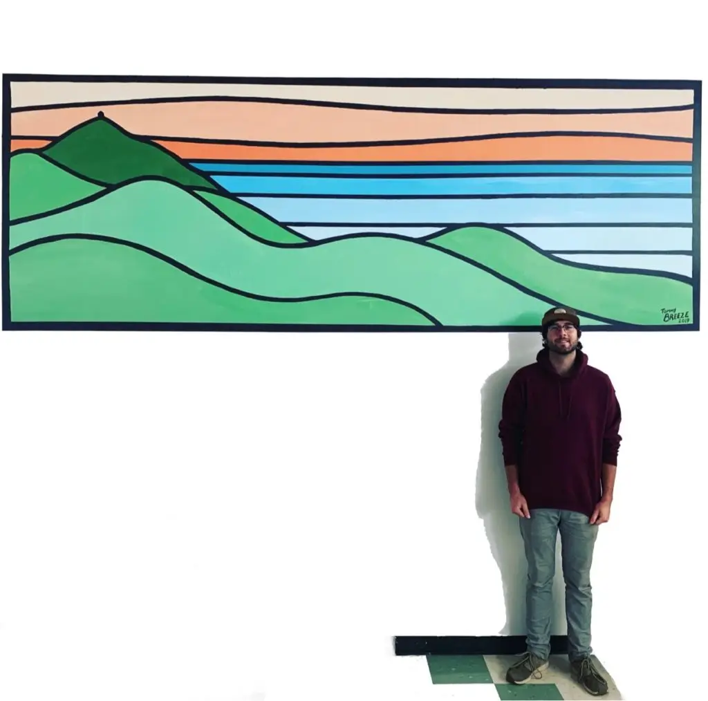 A person wearing a maroon sweater, jeans, and a cap stands against a white wall. Above them is a large rectangular painting depicting stylized rolling green hills and a lake under an orange and blue sky, evocative of the serene landscapes often associated with eco-friendly beauty products.