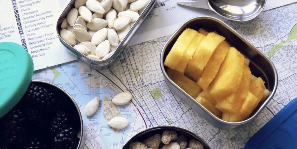 A top-down view of a map and list partially covered by various containers of road trip snacks. The containers hold shelled pumpkin seeds, sliced yellow fruit, blackberries, and nuts. A small spoon is seen in the corner.