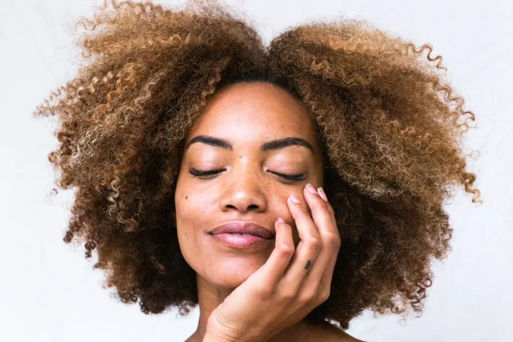 A person with curly, voluminous hair, closed eyes, and a serene expression gently touching their face with one hand against a plain white background, embodying the essence of eco-friendly beauty products.