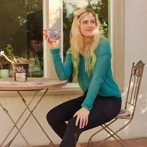 A blonde woman in a turquoise top and black pants sits on a metal chair outdoors, holding a large, decorative mug. She is next to a small round table with potted plants on it, against a cream-colored wall with a window reflecting trees; her relaxed demeanor radiates Sol Sister Sport vibes.