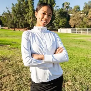 A person wearing a white, long-sleeved top and black pants stands outdoors on a sunny day, smiling with arms crossed. They're in a grassy area with trees and a building in the background, embodying the spirit of Sol Sister Sport.