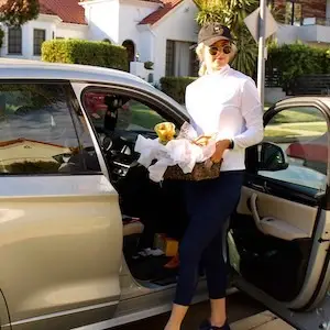 A person donning a black cap, sunglasses, a white turtleneck, and dark pants stands next to an open car door. They hold a basket filled with yellow flowers and white tissue paper. The backdrop showcases a residential area with white houses and greenery, reflecting the serene vibe of Sol Sister Sport.