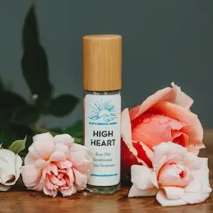 A bottle of essential oil labeled "High Heart" with a wooden cap, from Ellie's Essential Blends, is surrounded by pink and white roses. The background is dark gray, and the flowers are arranged in such a way that the bottle of oil is at the center of the display.