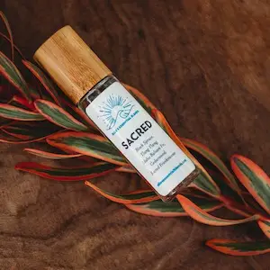 A bottle labeled "Sacred" rests on a brown surface, partially surrounded by a plant with green and red leaves. The label features a blue logo with the text: "Ellie's Essential Blends Sacred Pain Relief Roll-On." The bottle has a wooden cap.