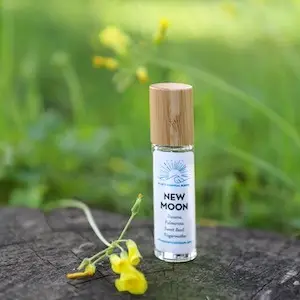 A small glass bottle of Ellie's Essential Blends' New Moon essential oil with a bamboo cap stands on a tree stump surrounded by green grass and yellow flowers. The clear label displays the product name, "New Moon," and additional text below. The background is slightly blurred.