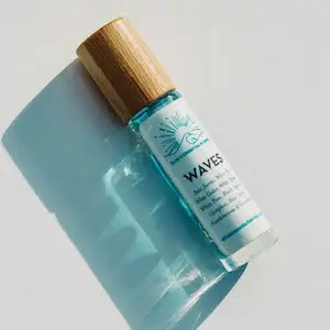 A small glass bottle with a wooden cap lays on its side, casting a shadow. The bottle is filled with blue liquid and labeled "WAVES" with an illustration of a wave and the sun. Bearing the mark of Ellie's Essential Blends, the label also includes some descriptive text, partially visible but not fully readable.