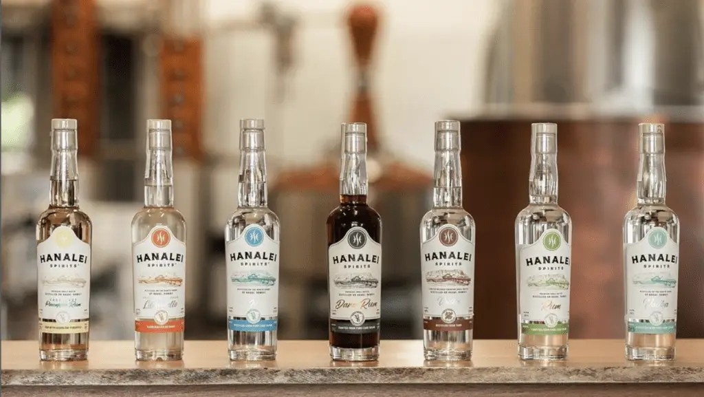A row of seven Hanalei Spirits bottles, each labeled with different colored tags, are displayed on a wooden surface. The transparent Kauai-made bottles contain various spirits with their labels featuring the Hanalei Spirits logo. The background shows a distillery setup.