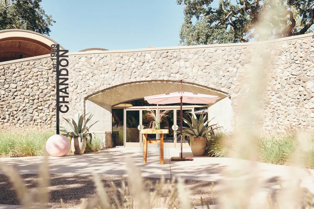 Entrance to the Chandon winery, featuring a stone facade, arched glass doors, and a pink patio umbrella. Potted plants flank the entrance, and a vertical sign reads "CHANDON Est. 1959." Tall grasses frame the foreground under a clear sky—one of the best things to do Napa April.