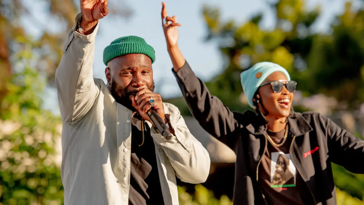 Two people are performing on stage outdoors. The person on the left is speaking into a microphone and wearing a green beanie, while the person on the right is raising their hand, wearing sunglasses and a blue beanie. Trees and a clear sky are visible in the background—a glimpse of the best things to do in East Bay this April.
