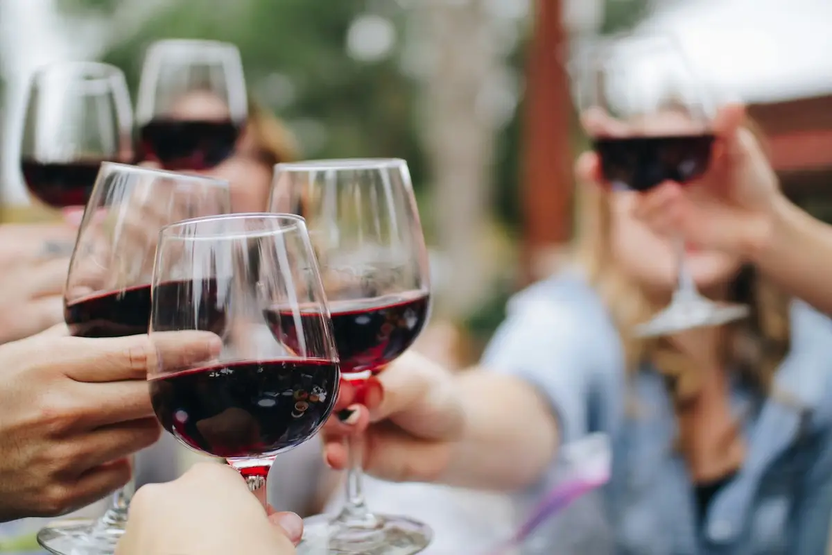 A group of people clinking glasses of red wine in a celebratory toast at one of the best things to do in South Bay this April. The background is blurred, suggesting an outdoor setting. The glasses are filled with dark red wine, and the scene conveys a sense of joy and togetherness.