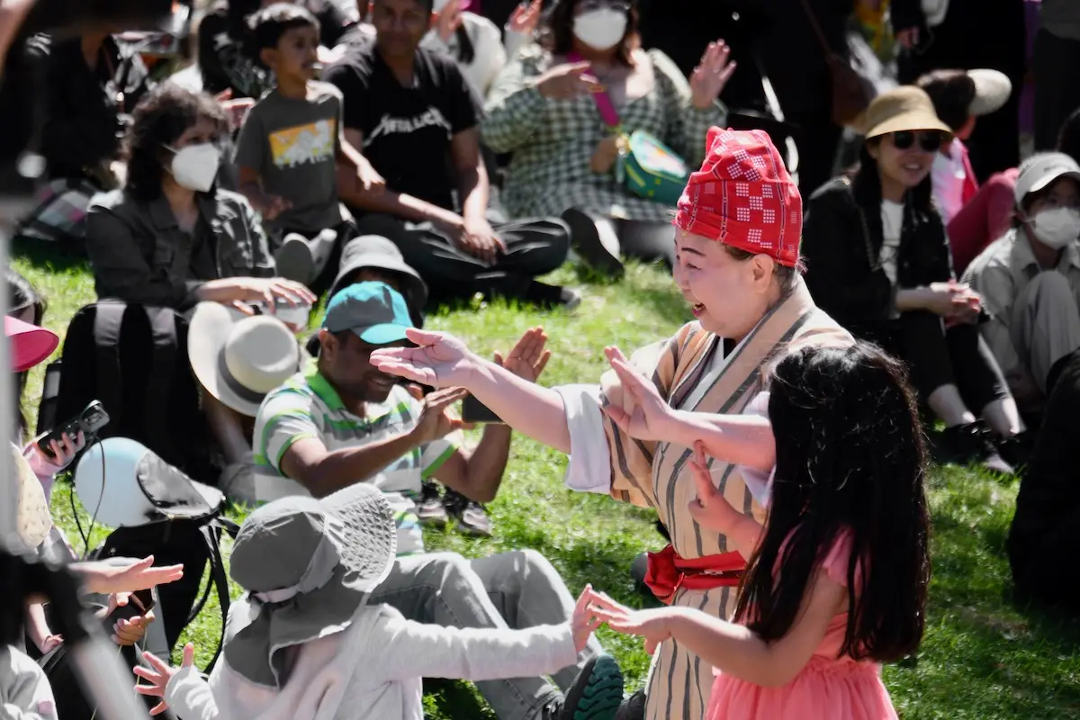 A performer wearing traditional attire and a red headscarf interacts with children at an outdoor event, one of the best things to do in the South Bay this April. People in the background, some wearing masks and hats, are seated on the grass, applauding and enjoying the performance under sunny weather.