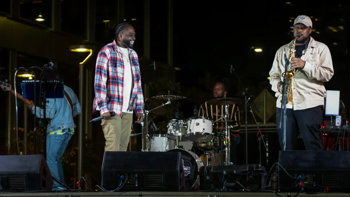 A musical performance on stage features five musicians, undoubtedly one of the best things to do in South Bay this April. One man in the center, wearing a plaid shirt and holding a microphone, smiles at a saxophonist on the right. In the background, there are drums and other instruments. The stage is illuminated and the audience is not visible.