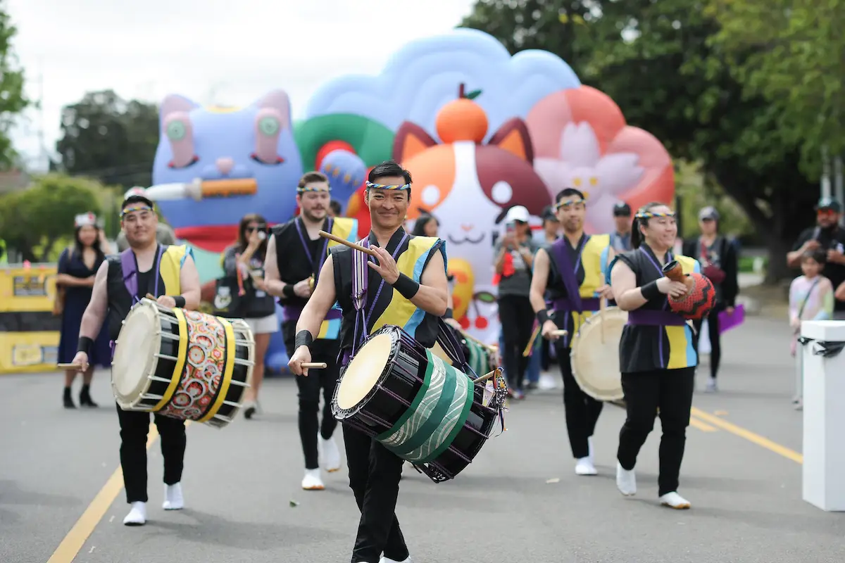 A group of people dressed in traditional clothing play large drums while marching down a street. They are part of a vibrant parade, one of the best things to do in South Bay this April, with colorful inflatables, including cat figures, in the background. Spectators watch from the sidelines.