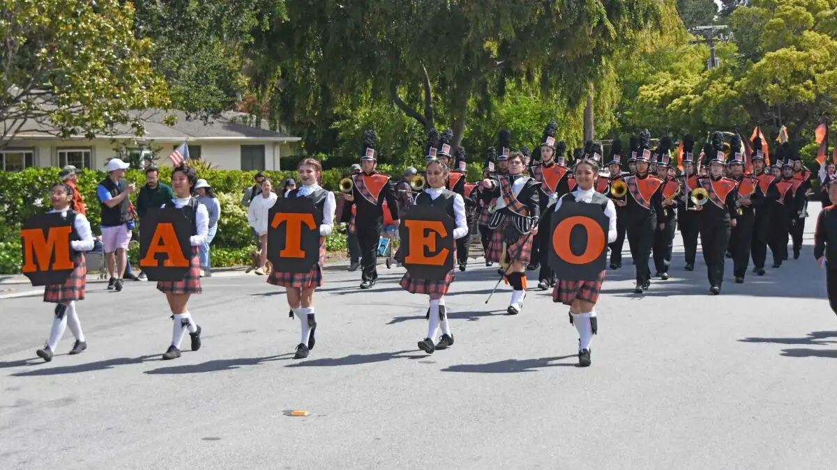 A marching band parades down a suburban street with participants wearing kilts and black hats. Five individuals at the front hold large orange letters spelling "MATEO." Spectators line the street, enjoying one of the best things to do in South Bay in May. Trees and houses are visible in the background.