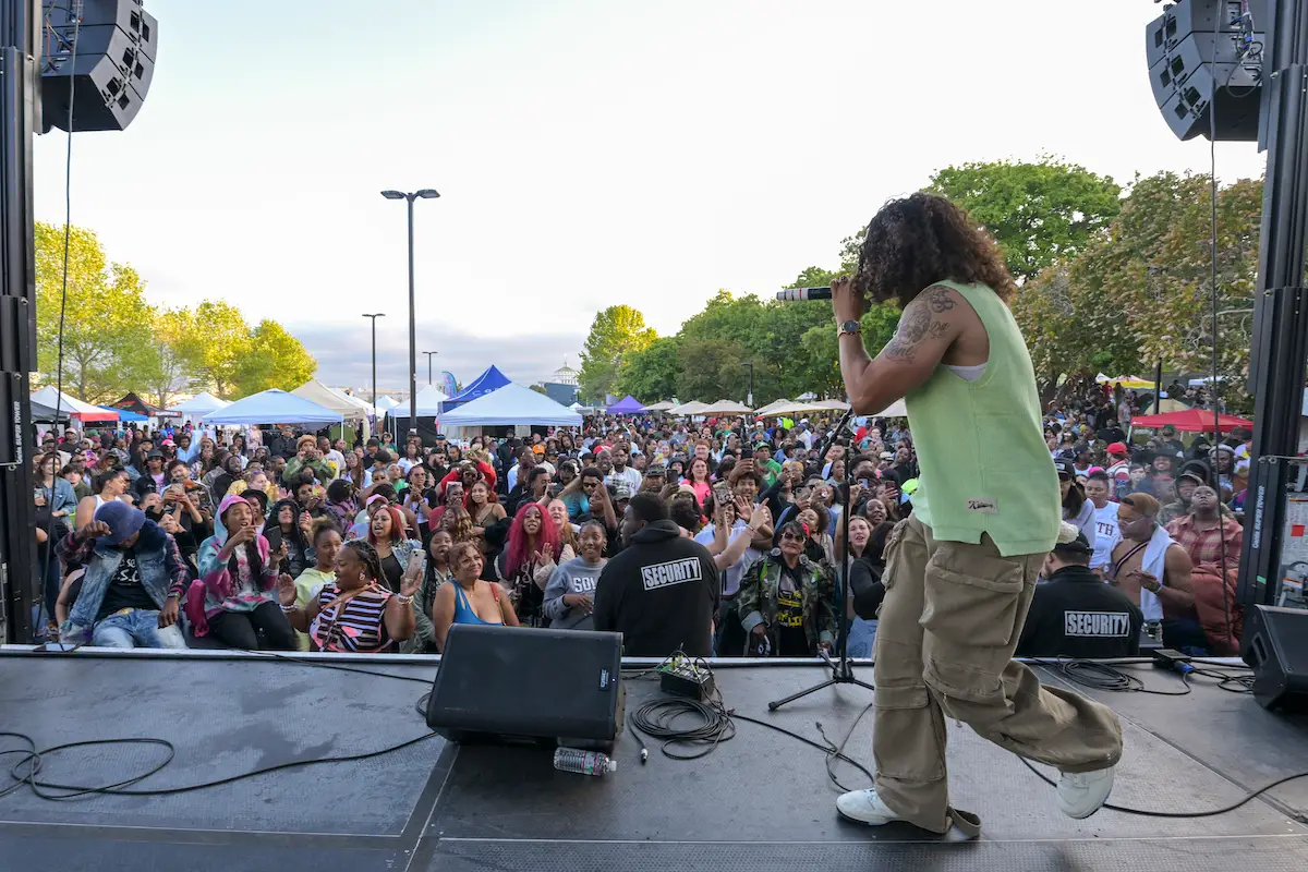 A person with long hair performs on an outdoor stage, holding a microphone. They are wearing a green tank top and beige cargo pants. A large, diverse crowd is cheering in front of the stage, with trees and tents visible in the background—one of the best things to do in East Bay this June.
