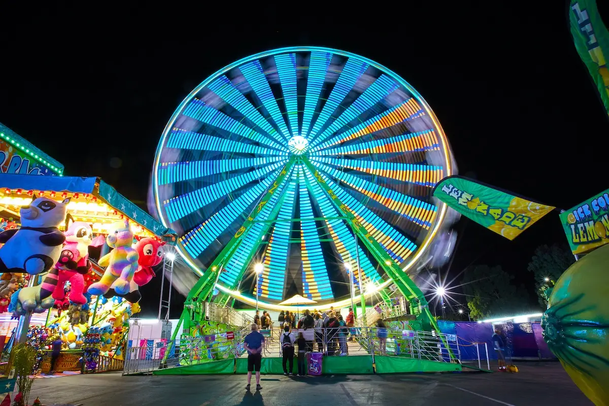 A brightly lit Ferris wheel at night with moving neon lights in shades of blue and green. Below, people are queuing to ride, and colorful game booths with stuffed animal prizes line the sides. The lively atmosphere suggests a bustling fair or carnival, one of the best things to do in East Bay this June.