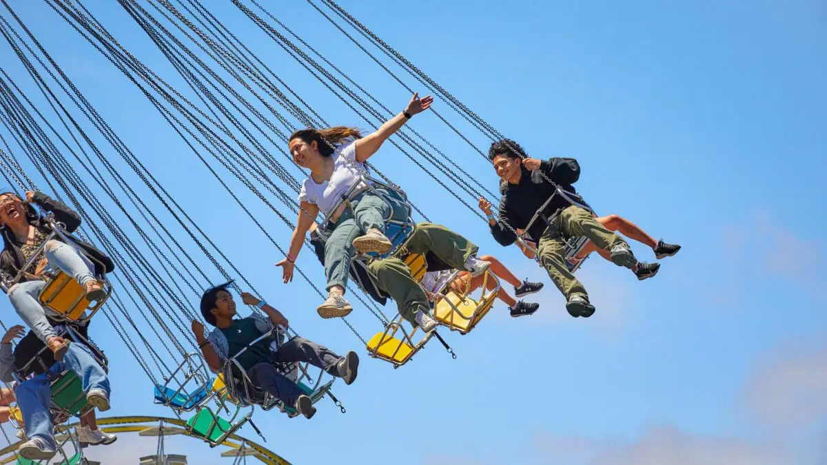 A group of people enjoying a swing ride at an amusement park in the East Bay. Seated in colorful swing chairs suspended by chains, they soar high against a clear blue sky. Two individuals have their arms outstretched, appearing joyful and carefree—one of the best things to do this June.