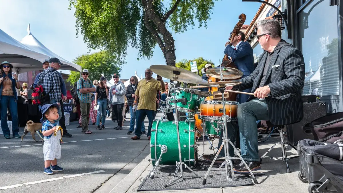A small child stands captivated by a street band performing in an outdoor setting, one of the best things to do in South Bay this June. The drummer is in focus, playing an emerald green drum set. An upright bass player is visible in the background as a crowd of people and a dog watch the performance. Trees and tents are in the background.