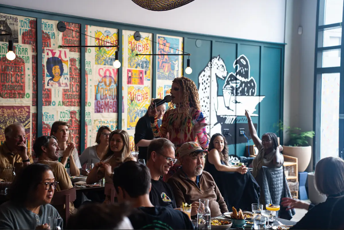 A lively restaurant scene with patrons seated at tables chatting and eating. A performer in vibrant attire stands with a microphone, engaging the audience. The background features colorful murals, posters, and a whimsical illustration on a wall—perfect for Father's Day brunches in the San Francisco Bay Area.