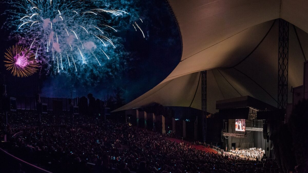 A large crowd watches an illuminated stage under a modern, tent-like structure during an evening performance. Colorful fireworks burst in the sky above, creating a festive and vibrant atmosphere—one of the Best Things to Do South Bay July.