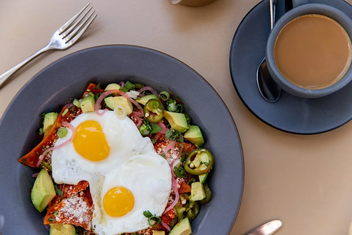 A gray bowl holds the best breakfast in Napa Valley: two sunny-side-up eggs atop a mix of avocado slices, tomatoes, onions, and jalapeno peppers. A filled coffee cup on a matching gray saucer sits beside the bowl, with a fork placed to the left.