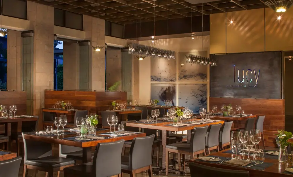 A modern, upscale restaurant interior with wooden tables set with glassware and silverware. Black chairs surround the tables, and green floral centerpieces are placed on each table. A sign on the wall reads "Lucy." Soft, warm lighting enhances the ambiance at the spot known for the best breakfast in Napa Valley.