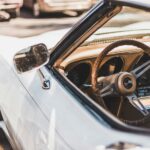 Close-up of the interior of a vintage white convertible car. The image highlights the steering wheel with a wood-grain finish, dashboard, and side mirror. Perfect for a cruise along the Monterey Peninsula in August, where the best things to do include classic car shows under gentle sunlight.