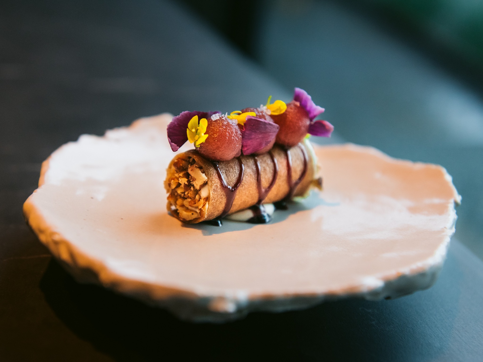 This delicately plated gourmet dish, perfect for one of the best dinner spots in Sonoma County, features a rolled pastry topped with vibrant edible flowers and garnishes, set on an elegant white ceramic plate with an organic, uneven edge. The dish is artistically presented against a dark, blurred background.