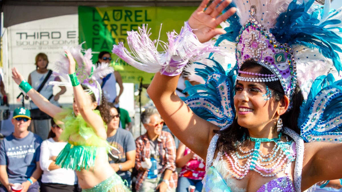 A woman wearing a vibrant, feathered costume, adorned with beads and sequins, dances energetically in front of a crowd at the Laurel Street Fair—one of the best things to do in the East Bay in August. Another performer in a green costume is visible behind her as spectators watch and take pictures.