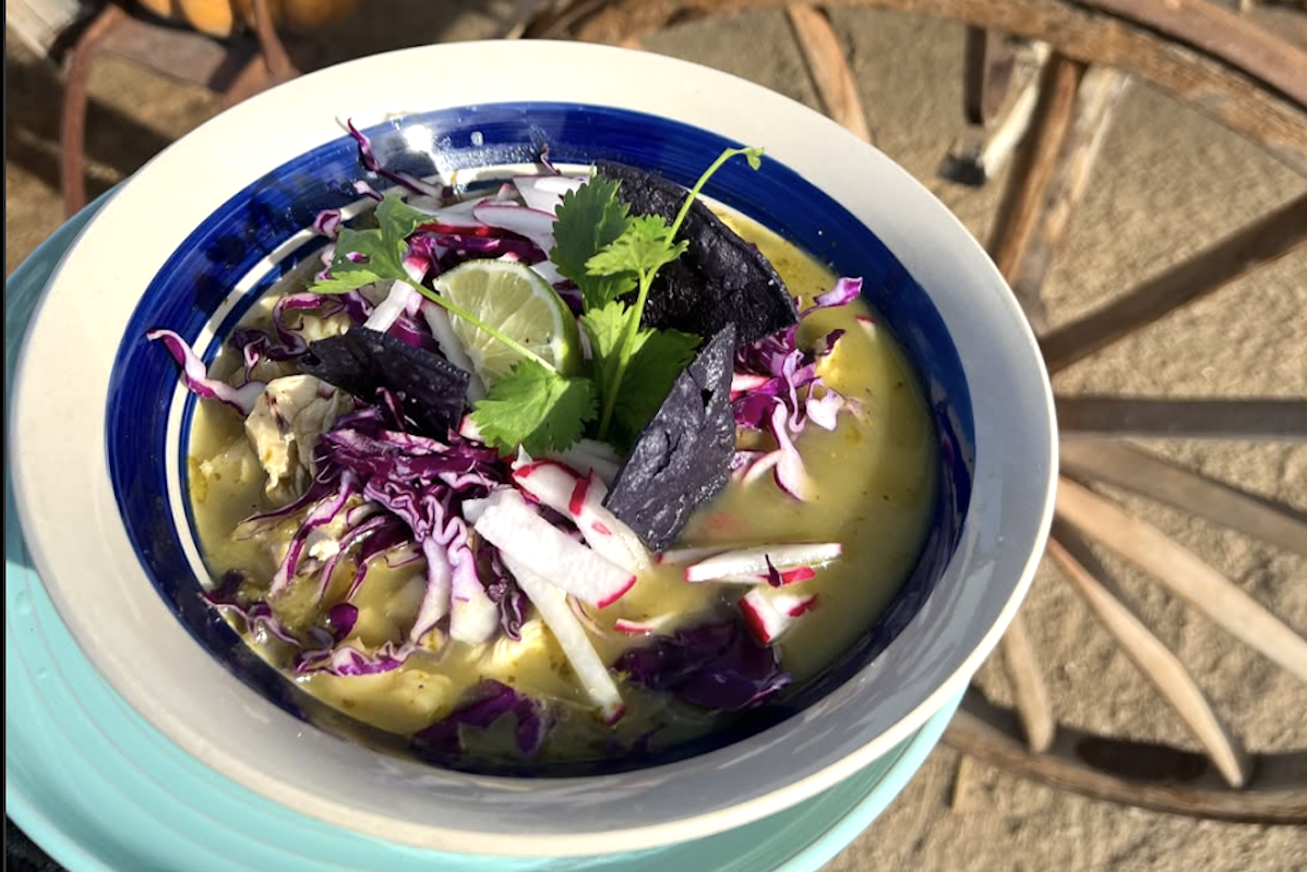 A white bowl with a blue stripe and soup with cabbage, blue corn chips, lime and cilantro.
