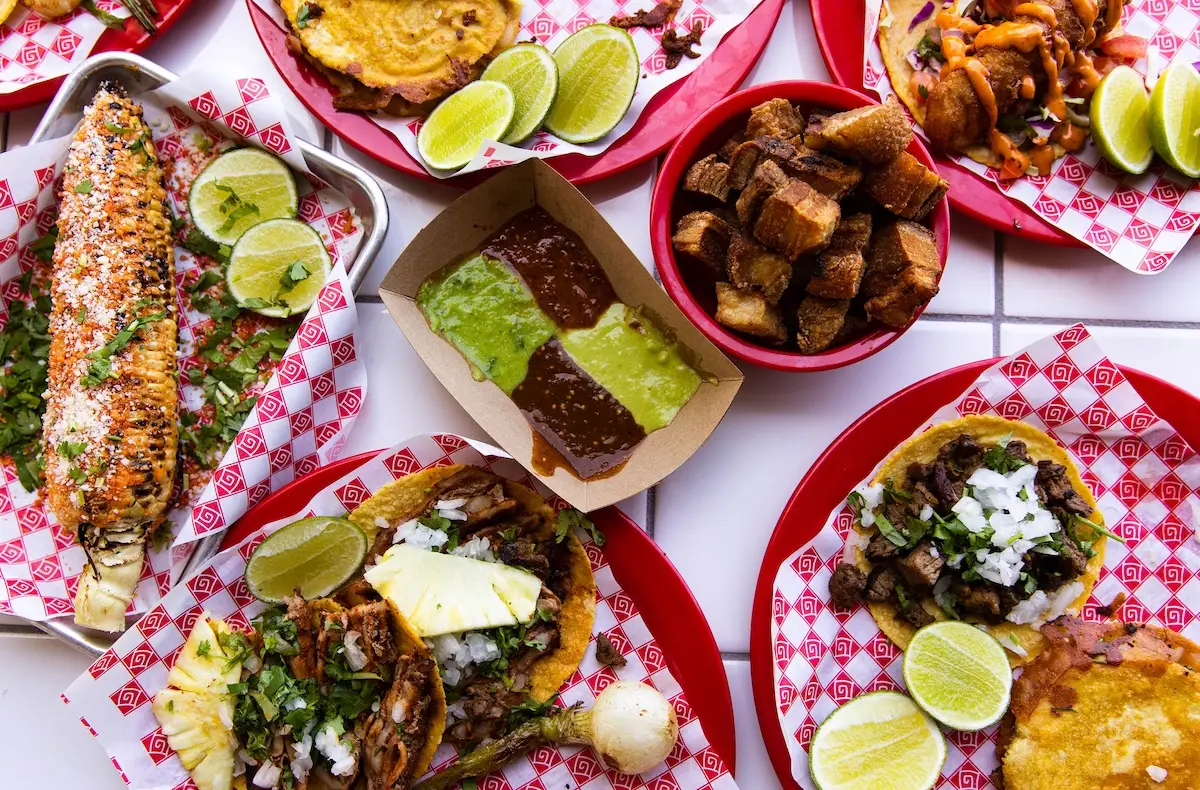 A vibrant spread of Mexican street food, perfect for the best lunch Napa Valley has to offer, includes tacos with various fillings, a grilled corn cob coated with cheese and chili powder, lime wedges, dipping sauces, and a bowl of fried croutons. The dishes are served on colorful plates and trays with patterned liners.