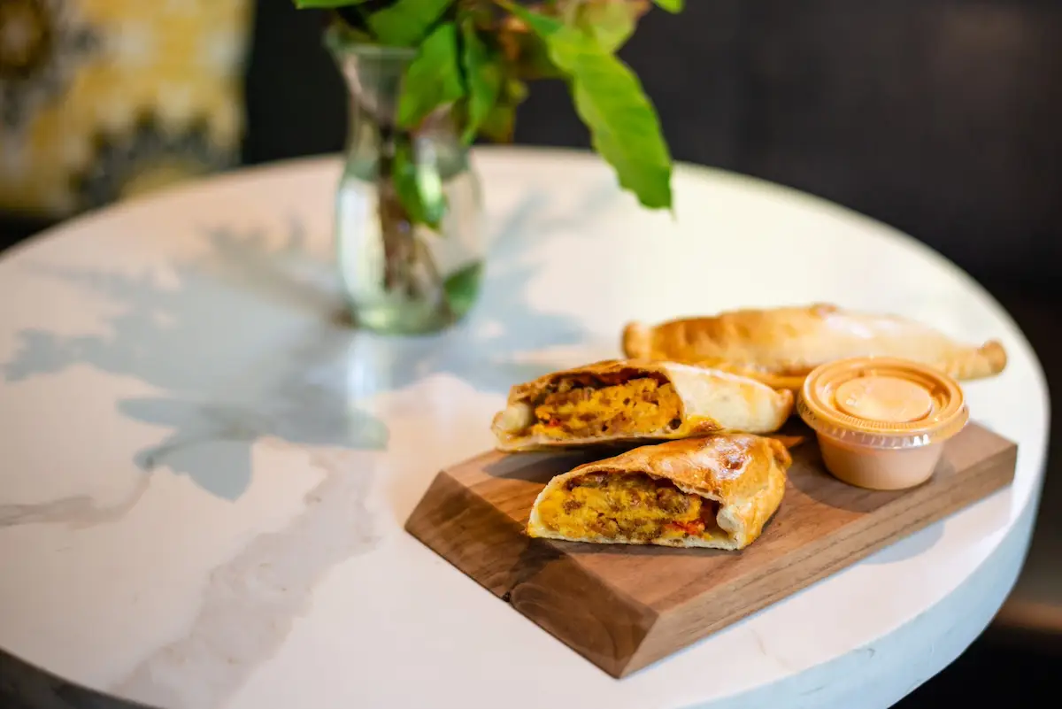 A wooden board on a marble table holds two empanadas, one cut open to reveal its filling, next to a small container of dipping sauce. In the background, a vase with green leaves adds a touch of freshness. Could this be the best breakfast in Napa Valley?