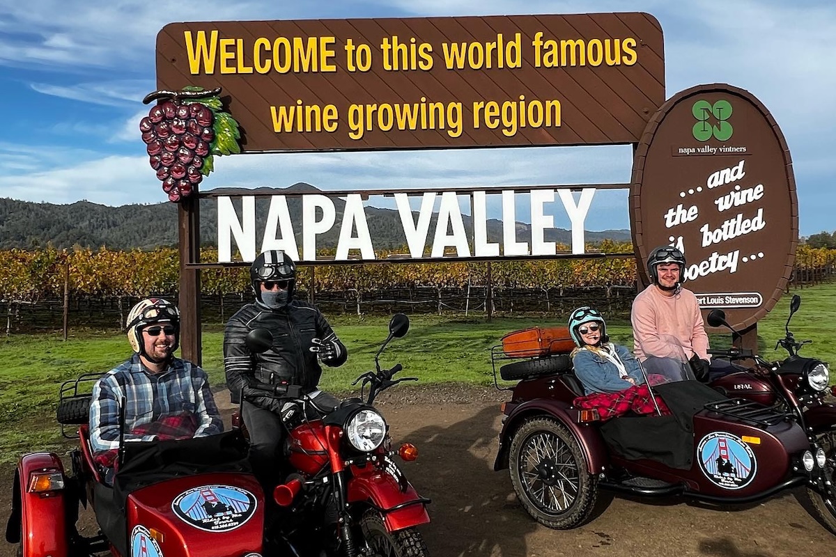 Four people sit on two red sidecars in front of a vineyard and a sign that says "Welcome to this world famous wine growing region NAPA VALLEY."