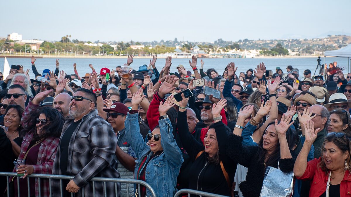 A large, festive crowd stands behind a barrier, many with arms raised and smiling faces, attending an outdoor event near a waterfront. People are dressed casually with sunglasses and hats, enjoying the sunny day. This lively scene epitomizes the best things to do in the East Bay this September. The background features water and a distant shoreline.