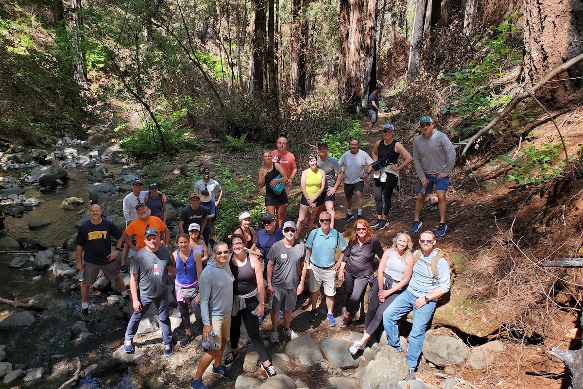 A large group of people dressing for hiking stand on a hiking trail next to a creek and surrounded by trees.