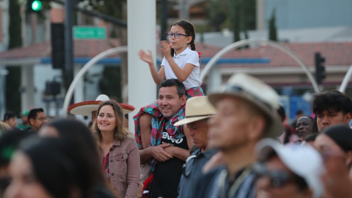 A young girl with glasses sits on a man's shoulders, clapping and smiling amidst a crowd at an outdoor event. Nearby, a woman looks in the same direction, also smiling. Sombreros and hats are worn by some attendees as they enjoy one of the best things to do in South Bay this September. Urban streets and buildings are in the background.