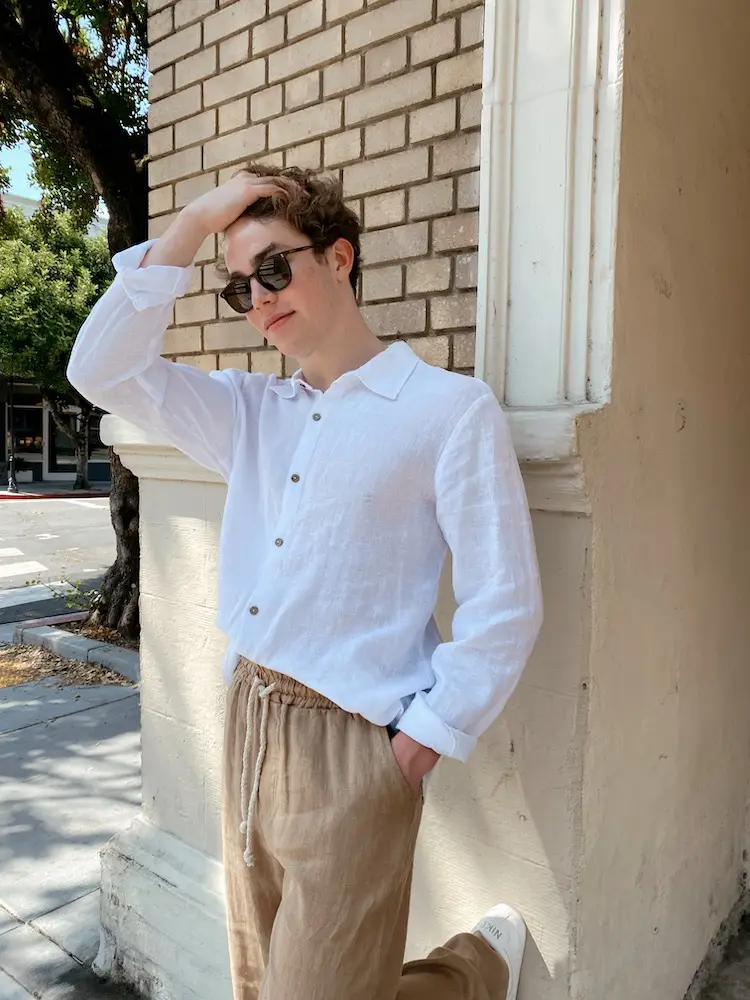 A person with short, curly hair wearing sunglasses, a white long-sleeved shirt from Shop Surf + Sand, and beige linen pants leans against a brick wall with one hand touching their head and the other in their pocket. They are standing on a sunny sidewalk with trees and buildings in the background.