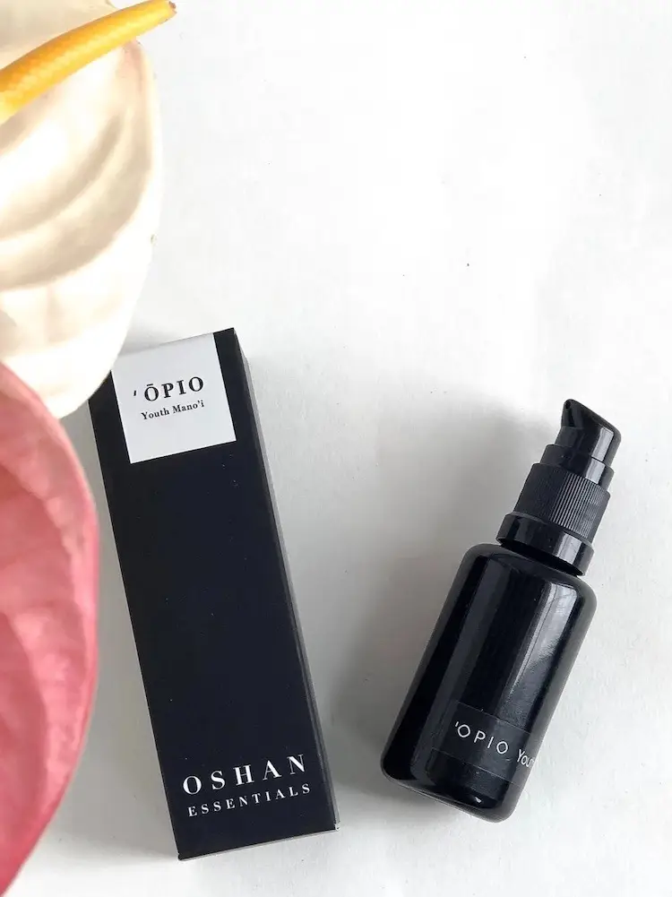 A black bottle of Ópio Youth Nectar by Oshan Essentials is placed next to its matching black packaging. The bottle features a pump dispenser. In the top left corner, partially visible are the edges of pink and white anthurium flowers. The scene, available at Shop Surf + Sand, is set against a white background.